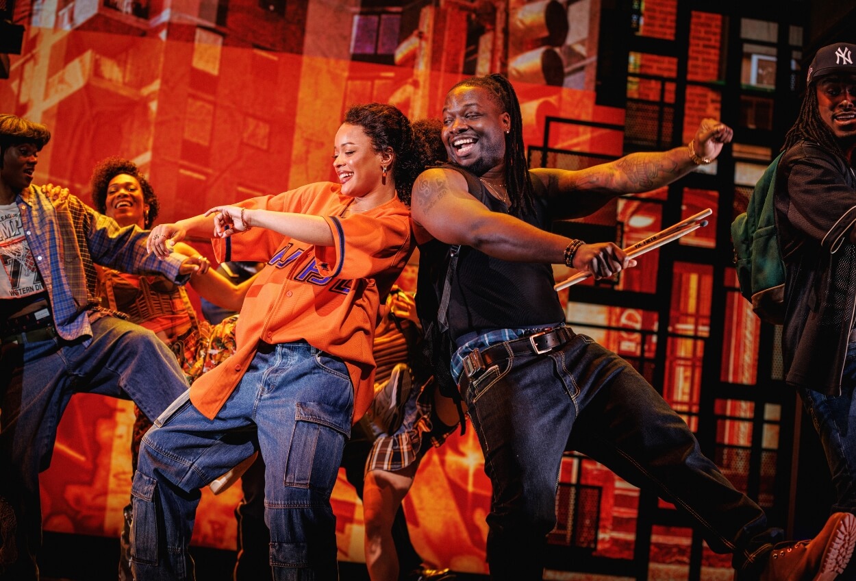 Two actors perform dramatically on a theater stage set to look like a colorful subway, conveying motion and emotion while one actor aims a prop gun.