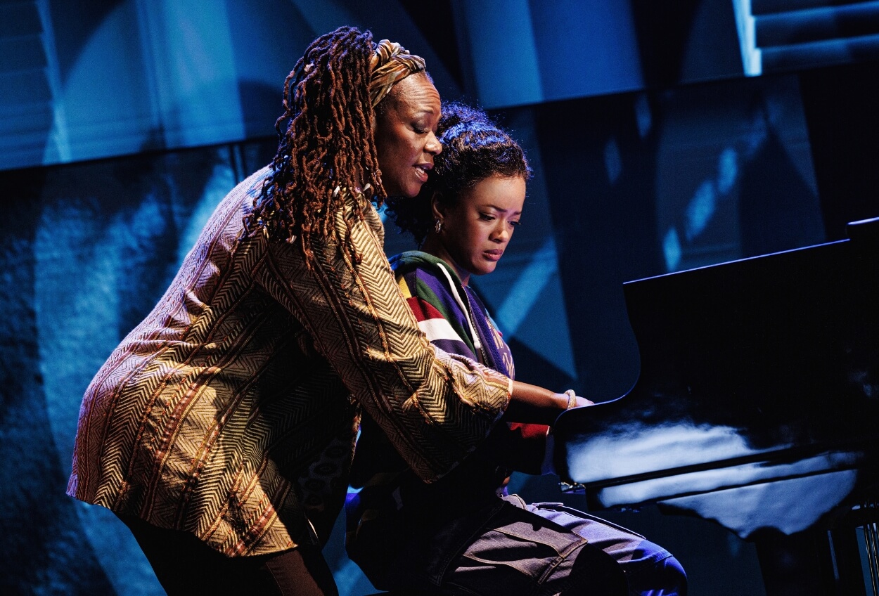 Two people at a piano, one teaching the other how to play, in a dimly lit stage setting with blue lighting. One person has long, curly hair, and the other has short hair.